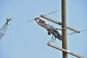 A lineworker in jeans, white shirt and a hardhat leans sideways off of a power pole to make repairs.
