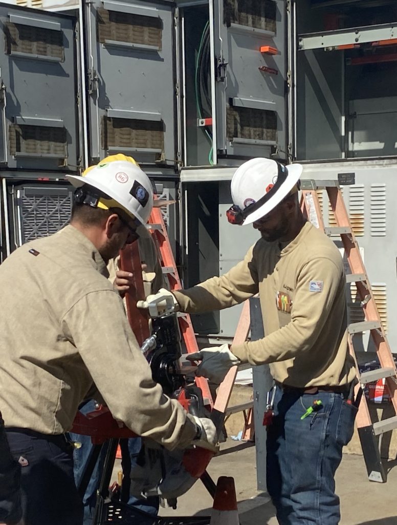 Image of two crew members wearing white constructions hats and beige long sleeve shirts assisting with an electrical repair.