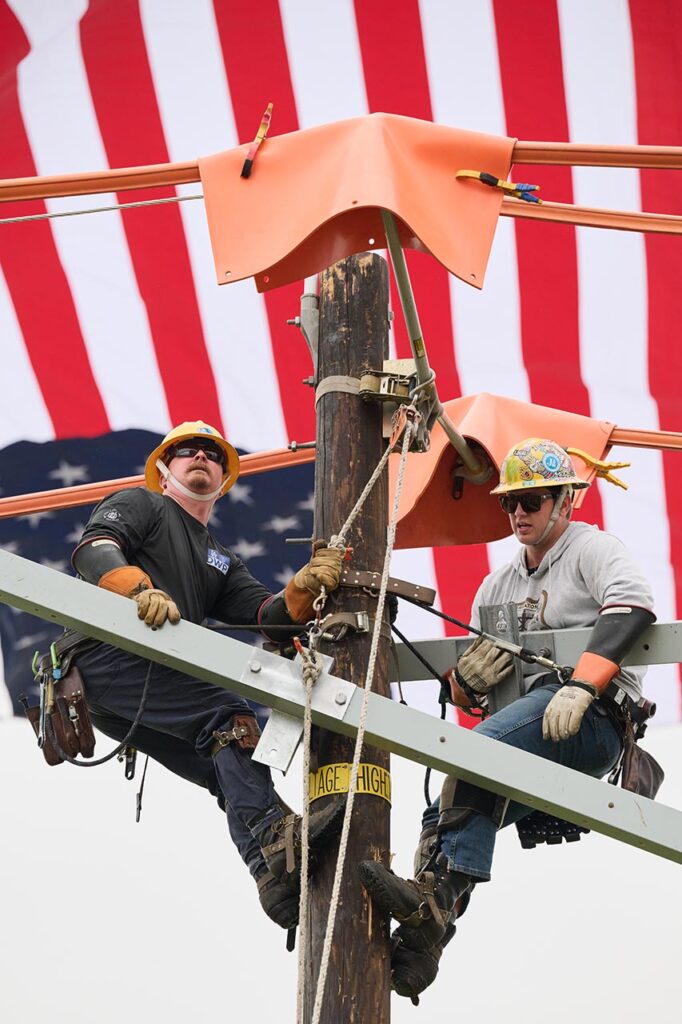 Two linemen atop an electrical power pole raise a cross arm via a pulley as they compete in the 2025 Lineman's Rodeo. Their backdrop is a large American flag.