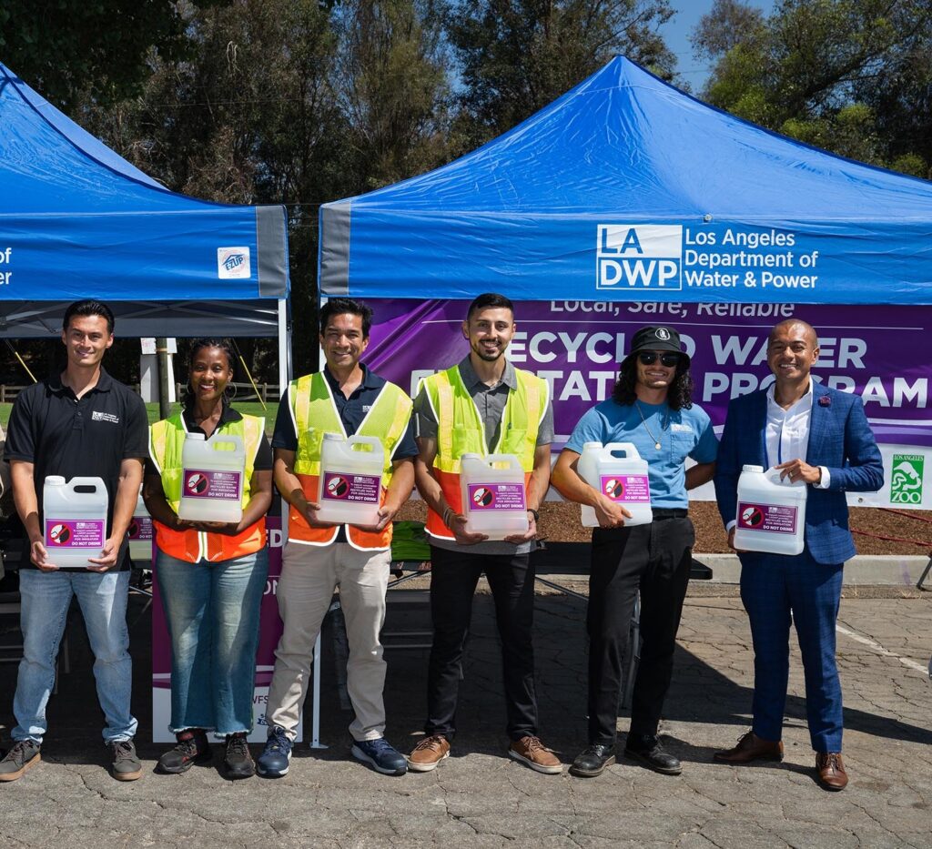 Group photo of diverse, smiling LADWP employees, five men and one woman. Three of them are wearing yellow/orange safety vests. Each one is holding a plastic jug of water with a purple label. They are standing in front of two, blue-colored easy ups with the LADWP logo.