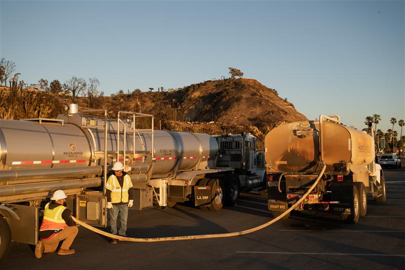 two field crew in high visibility neon vests with hard hats standing in front of large water tankers connecting a water hose with the sun setting in the hillside background 