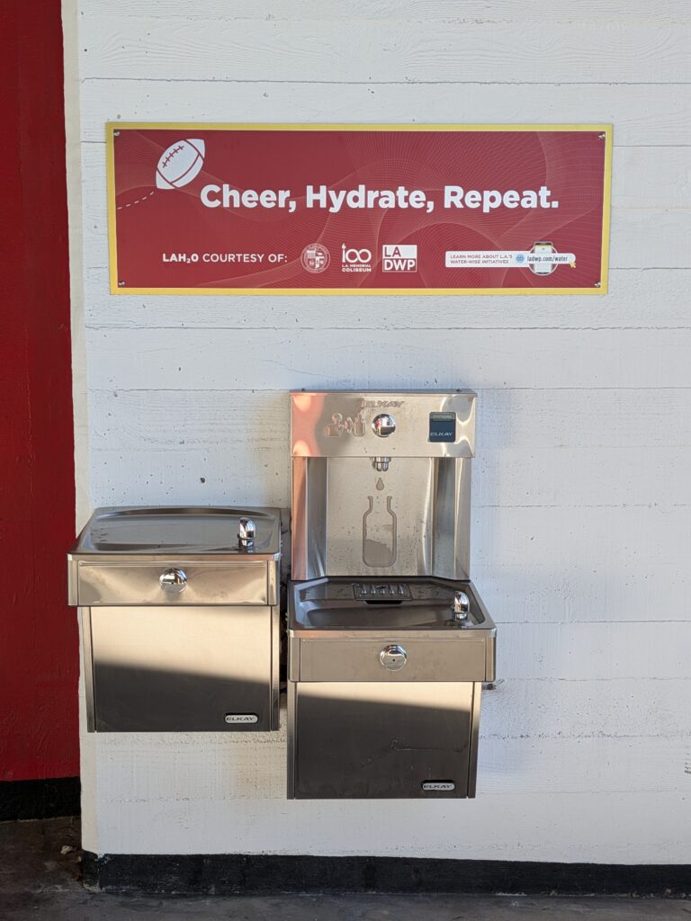 Shiny silver square water fountain affixed to a white concrete wall. Next to it is an equally shiny, silver, reusable bottle filler located at the L.A. Coliseum. A red colored sign with a thin gold border above it reads "Cheer, Hydrate, Repeat" in white lettering. A white football accompanies the text.
