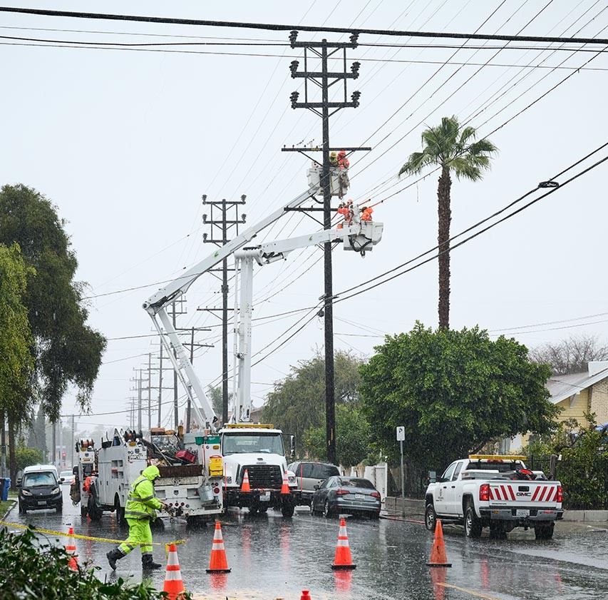 A field crew member wearing high visibility waterproof field gear walking on a street among several orange and white safety cones amidst a downpour of rain. Several other field crew members also wearing high visibility waterproof field clothing are in power buckets attached to a bucket truck high up in the power pole making repairs in the rain.