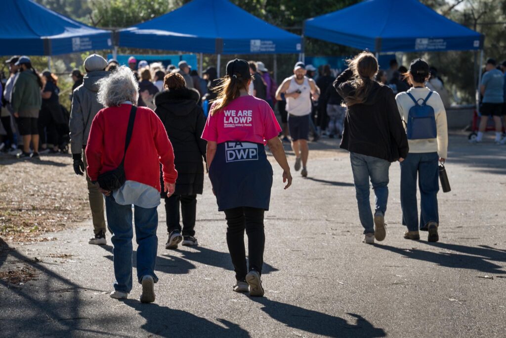 Participants walk the Hollywood Reservoir path as part of LADWP's Reservoir Walk celebrating Women's History Month and the 101st Anniversary of the Mulholland Dam. LADWP exhibitor booths draped in blue canopies are seen in the background.
