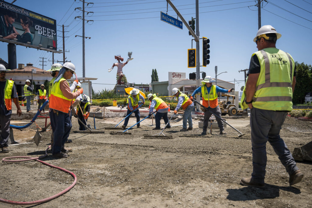 Crew members wearing hard hats and high-visibility vests perform maintenance on a street.