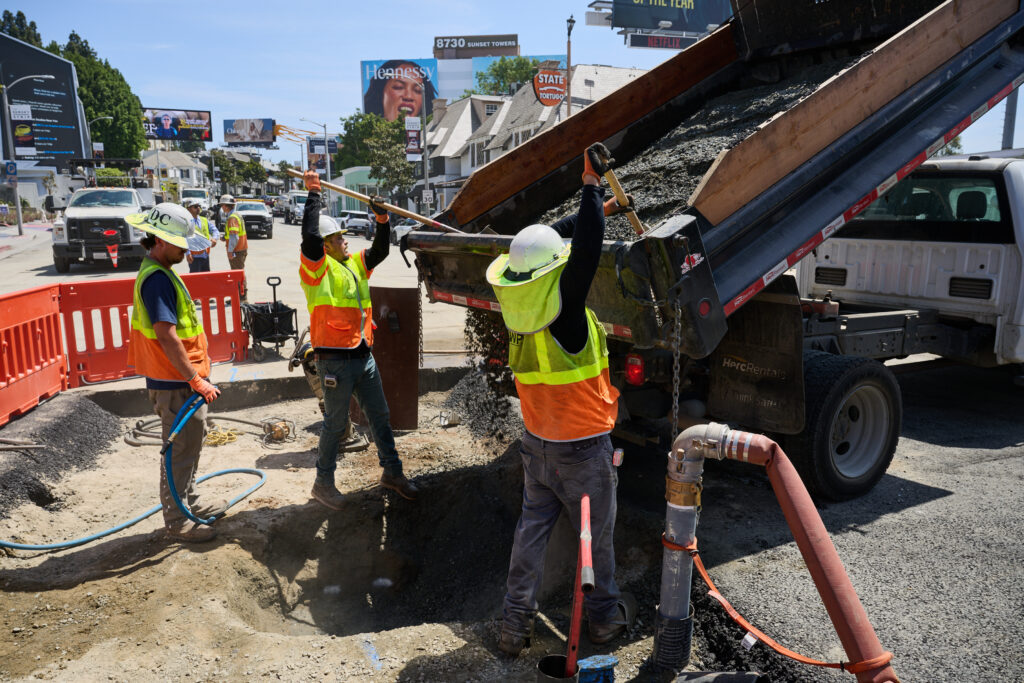 A dump truck dumps material into a worksite. Workers in hard hats and vests use shovels to help guide the material out.