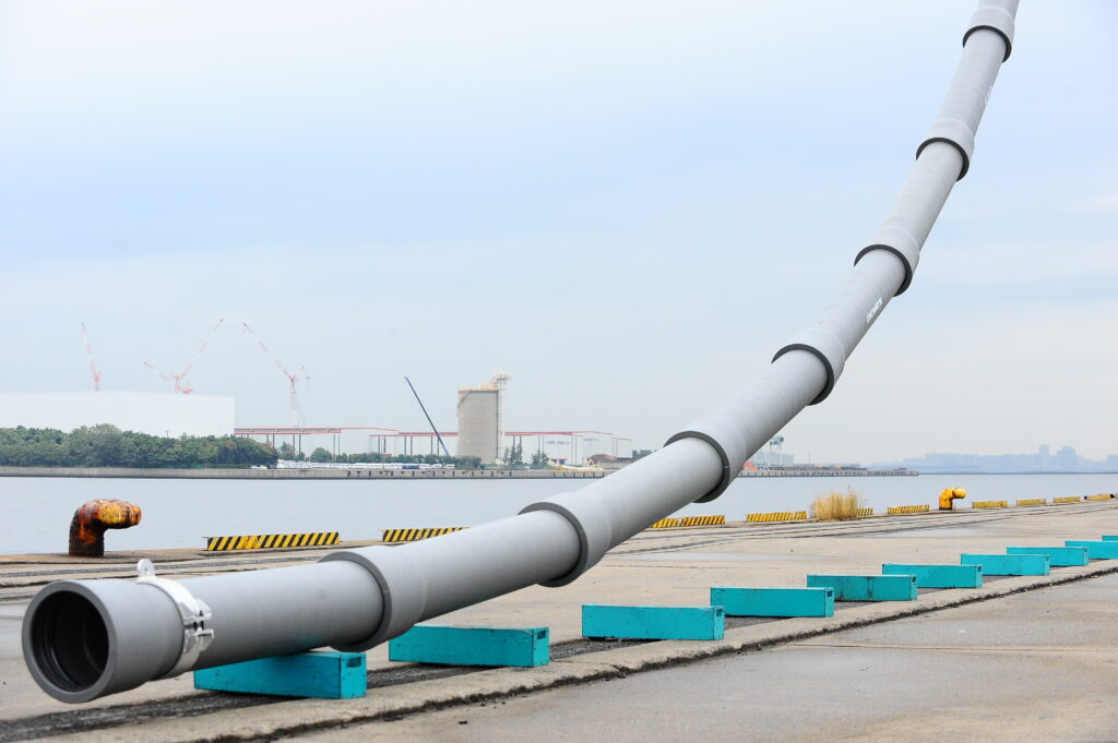 Large gray industrial pipeline curves across a waterfront loading area, with turquoise barriers, cranes, and a hazy city skyline under blue skies.
