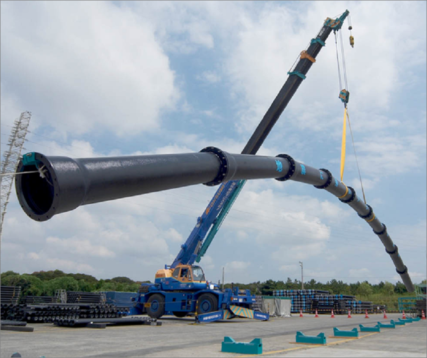 A mobile crane lifts a long, curved black pipeline section above an industrial yard, with stacked pipes, equipment, and a partly cloudy sky.