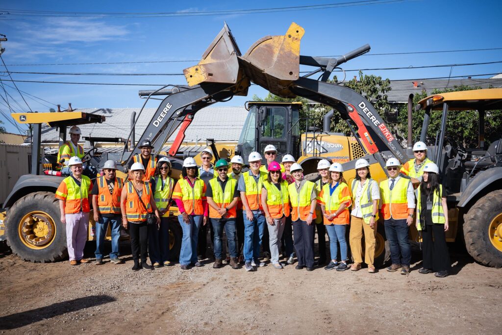 A group of people wearing hard hats and safety vests pose in front of construction equipment at the LADWP La Kretz Center groundbreaking ceremony